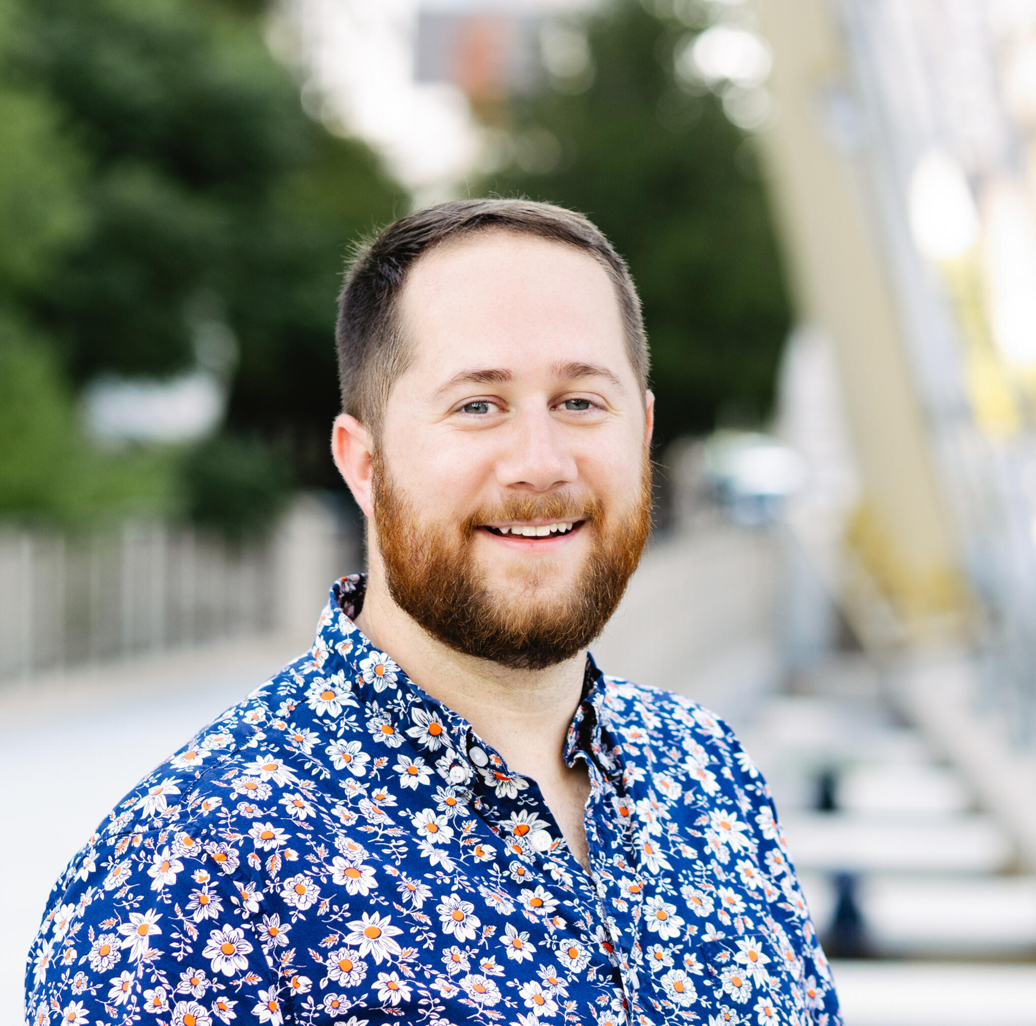 Headshot of Kevin Wedeen-Zittle in a floral shirt on a dark blue background outside.